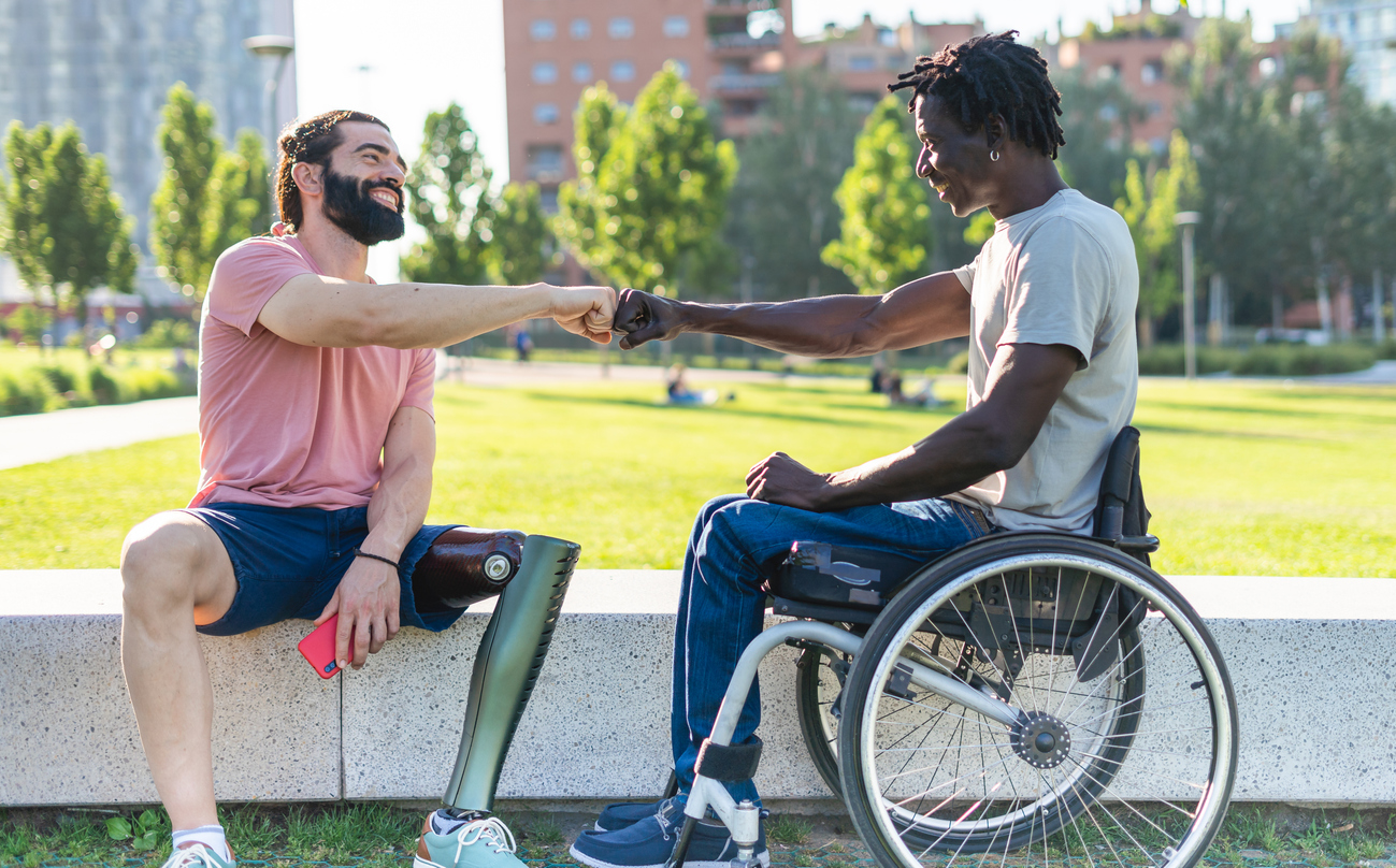 Photo of two men, one in a wheelchair and one with a prosthetic leg, giving a fistbump.
