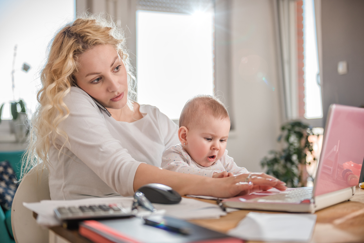 Woman holding baby on her lap, talking on smart phone at home office and using laptop.