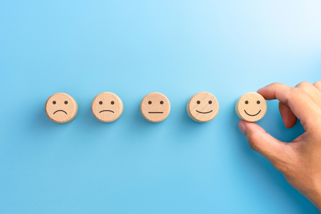 Five wooden faces from sad to happy. Hand reaching for the happy face.