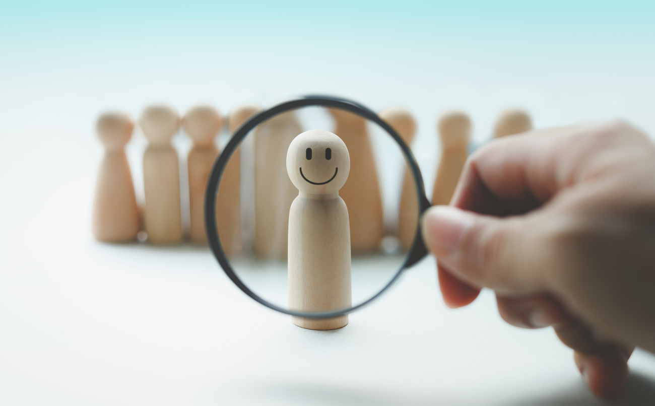 hand holding a magnifying glass over wooden figures, one with a happy face.
