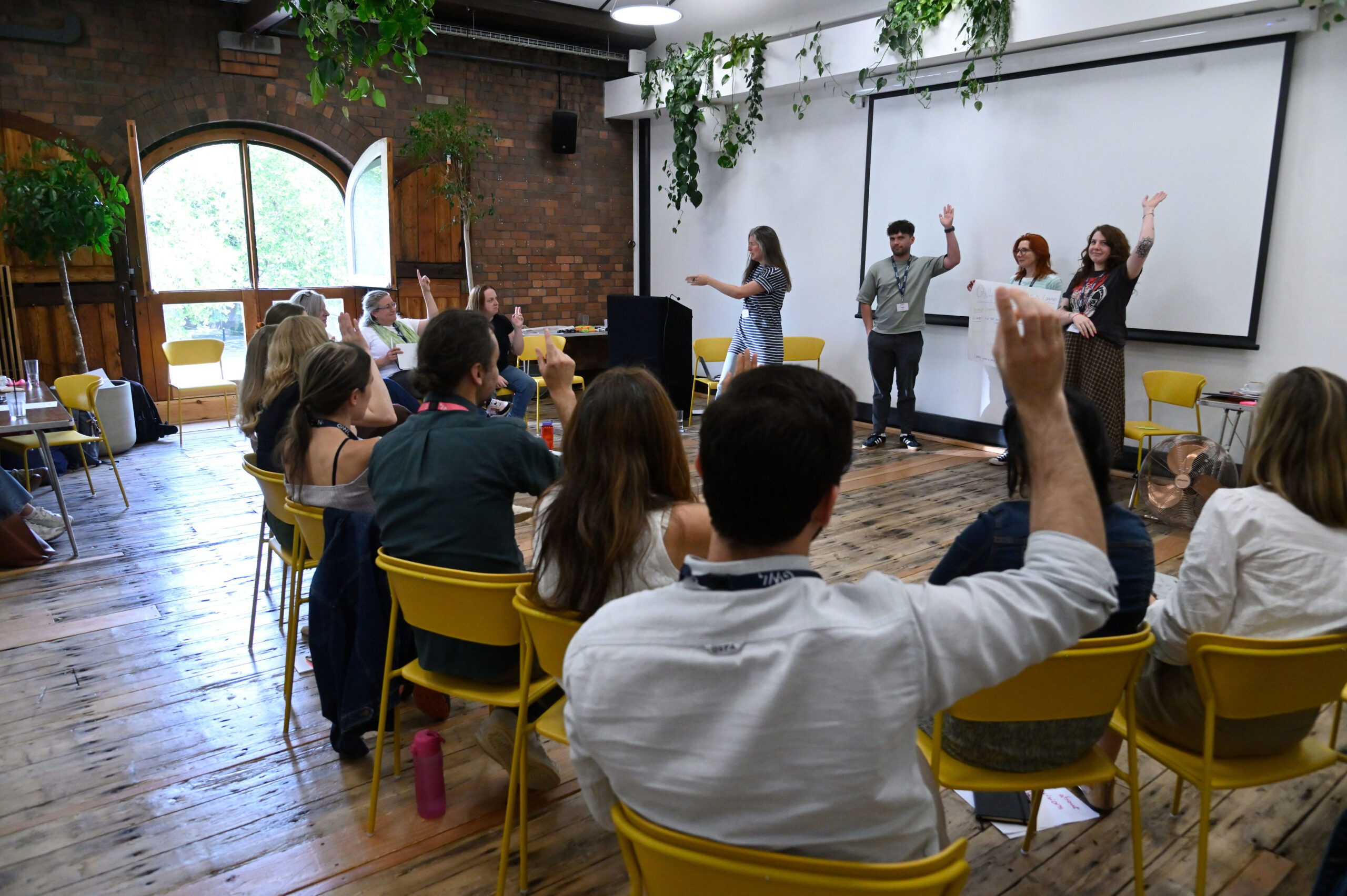 A large room with participants sat in a horseshoe shape and one group stood at the front pitching ideas.